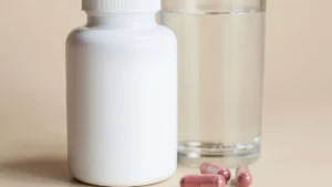 White supplement bottle next to a glass of water, with a few capsules on the table